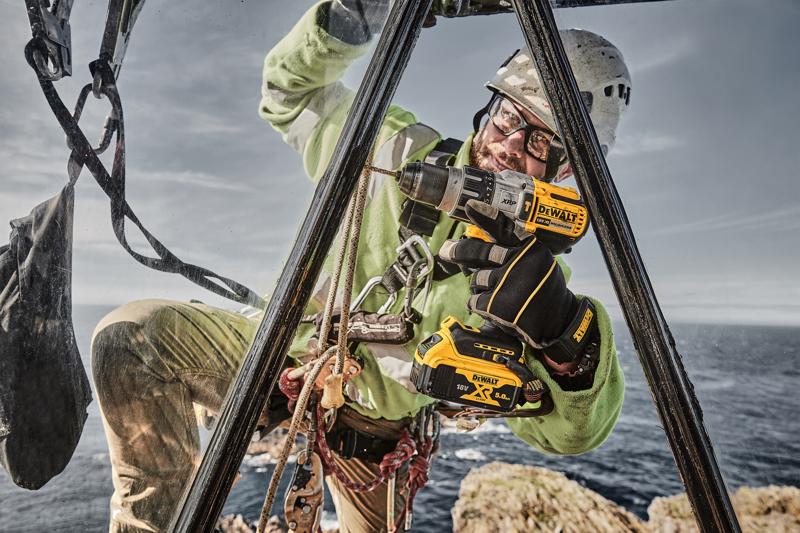 A worker wearing safety gear and gloves is using a DEWALT DCD996P2 cordless drill outdoors, performing maintenance or installation tasks on a metal structure above rocky terrain near the sea.
