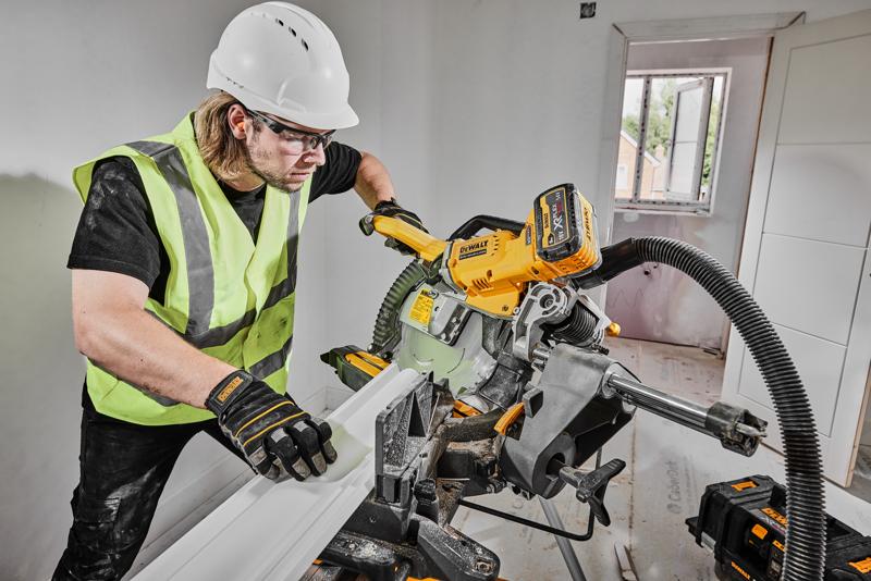A person wearing a safety vest, gloves, and hard hat is using a DEWALT DCS781X2 cordless miter saw to cut a piece of trim inside a room under construction. The saw is connected to a dust extraction system, and there are DEWALT tool batteries visible nearby.