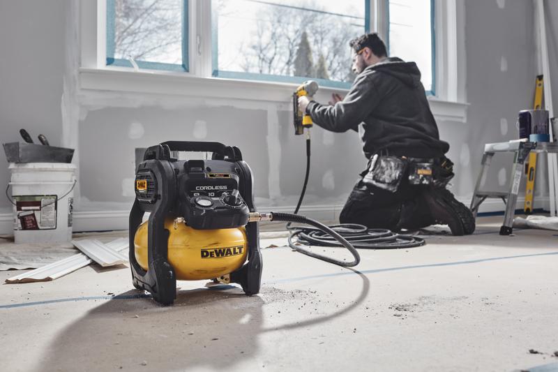A DEWALT DCC1018N cordless air compressor is placed on the floor of a room under renovation, with a worker using a pneumatic tool in the background near a window.
