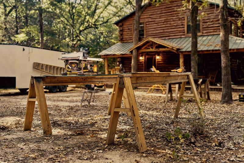 A DEWALT DWS780 miter saw placed on a wooden workbench outdoors, with a rustic log cabin in the background surrounded by trees and autumn leaves.