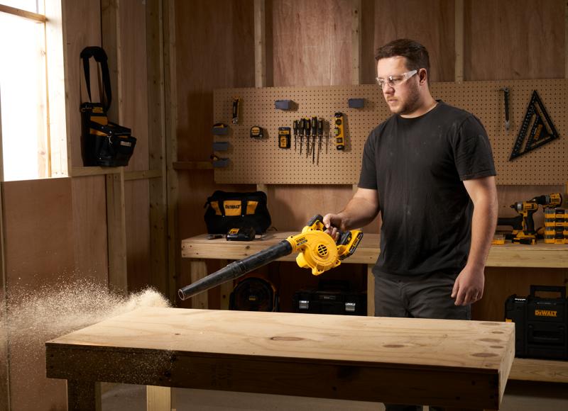A person using a yellow DEWALT DCV100 blower to clean sawdust off a wooden workbench in a workshop. The background shows various DEWALT tools and tool bags mounted on a pegboard and work surfaces.