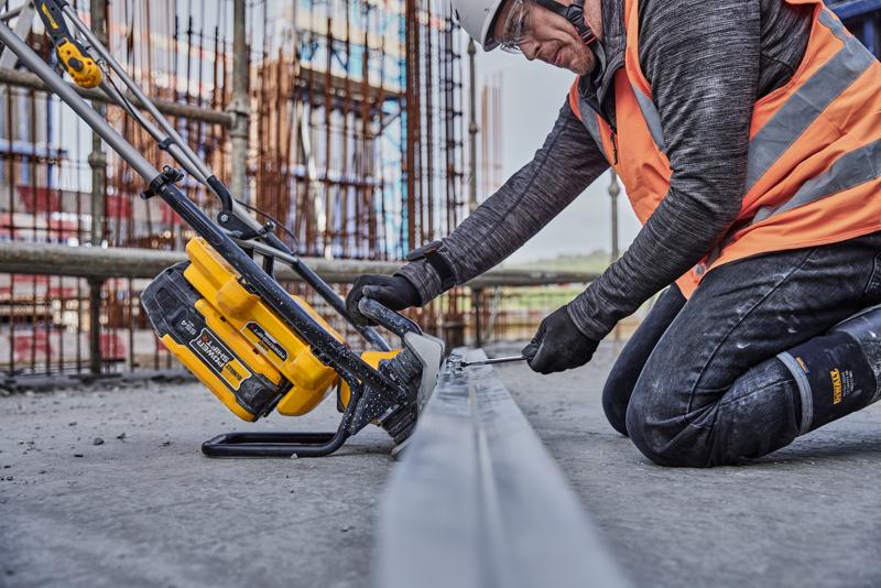 A person wearing safety gear is kneeling on a construction site, using a DEWALT cordless power tool on a metal beam. The surroundings show construction materials and unfinished structures.