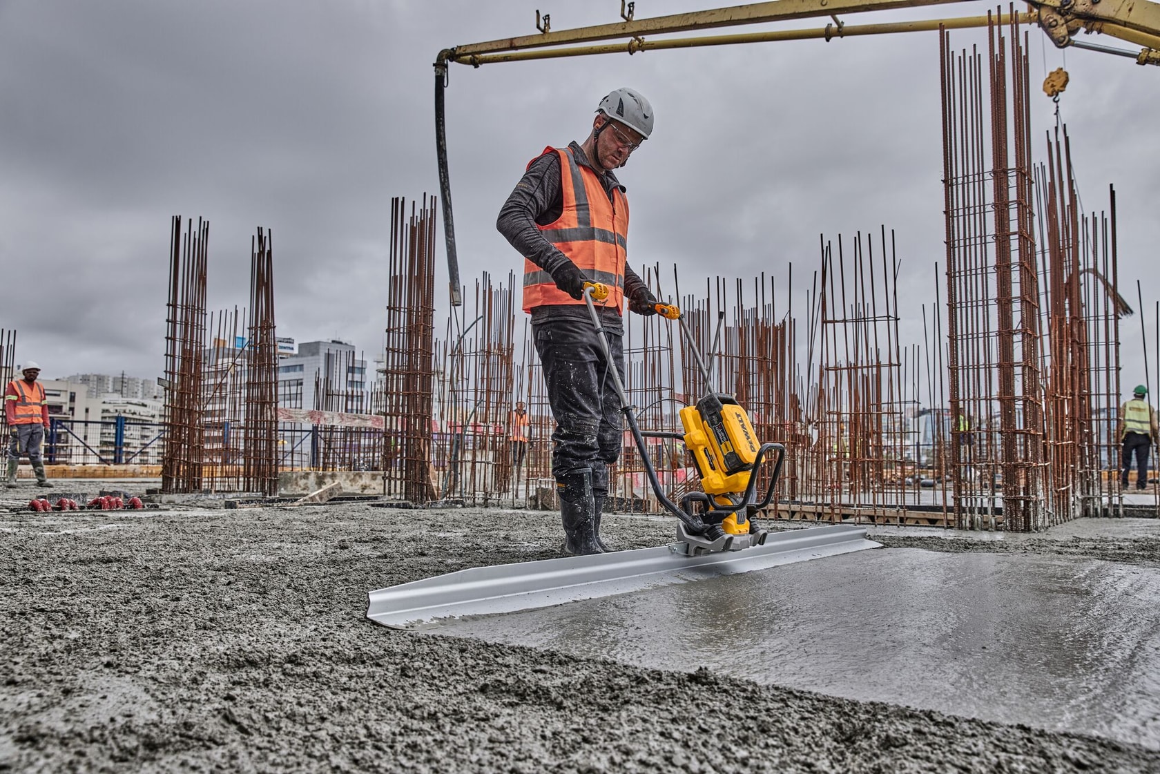 Worker uses a DEWALT power tool to smooth concrete at a construction site; steel rods are visible in the background.